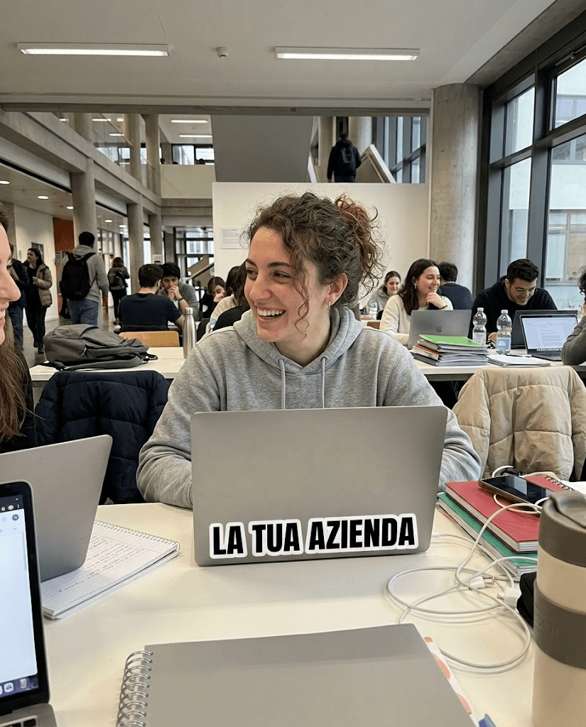 Una studentessa sorride con lo sticker La tua azienda sul laptop in un’aula studio universitaria
